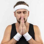 peaceful young handsome sporty man wearing headband wristbands putting hands together pray gesture with closed eyes isolated white space